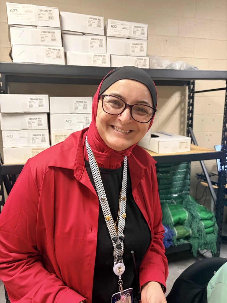 Woman in red jacket and glasses smiles in a room. Behind her, a shelf holds white boxes.