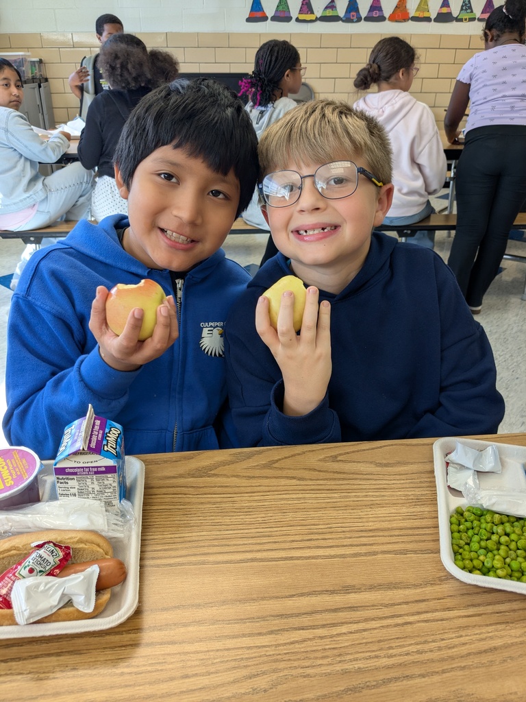 Two boys, one holding an apple and the other holding a banana, sitting at a cafeteria table.