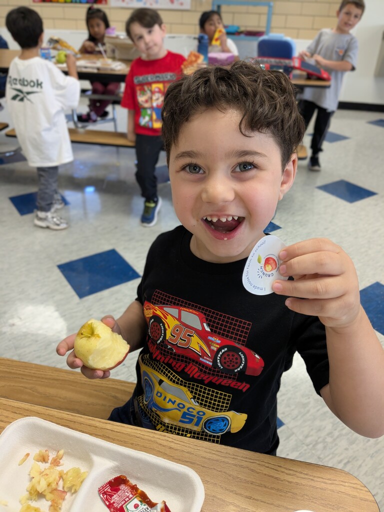 A child smiles in a classroom, holding a peeled apple and a white sticker. Other kids are seated at tables.