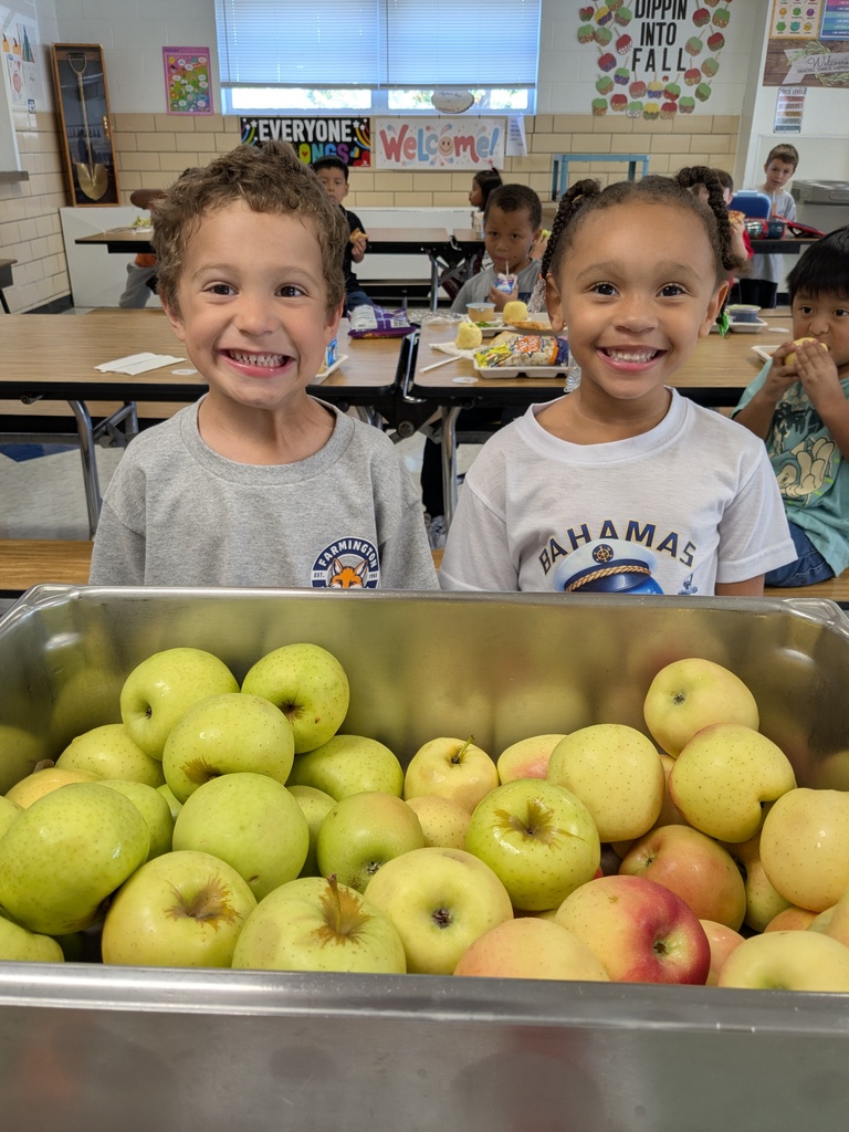 Two young children stand next to a bin filled with apples. The background shows a classroom with tables and chairs.