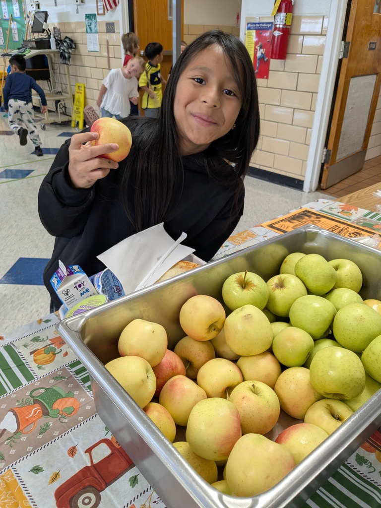 A girl in a black top holds an apple while smiling. A tray of apples is on a table.