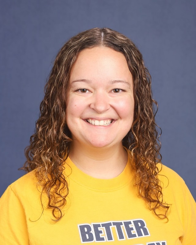 Woman with curly hair, wearing a yellow shirt. The shirt has the word "BETTER" printed on it.
