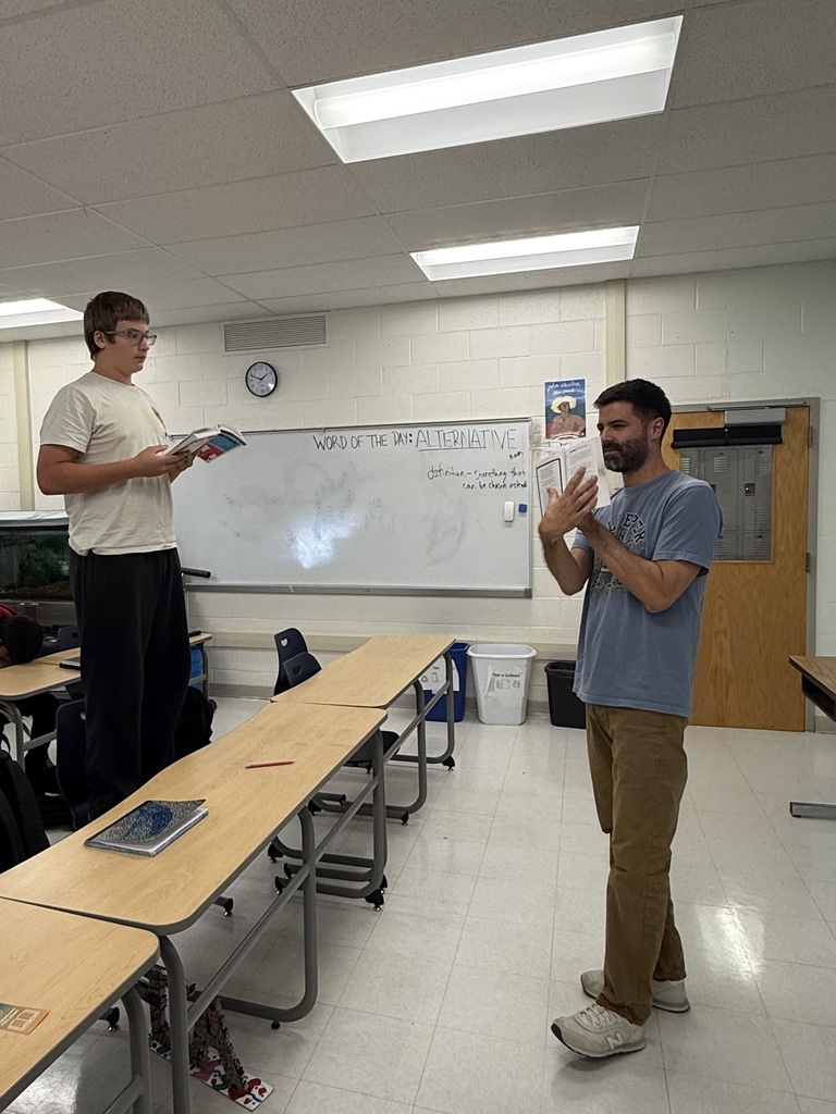 Two men in a classroom with white walls and ceiling lights, one reading a book and the other gesturing. Desks, chairs, and a whiteboard are in the background.