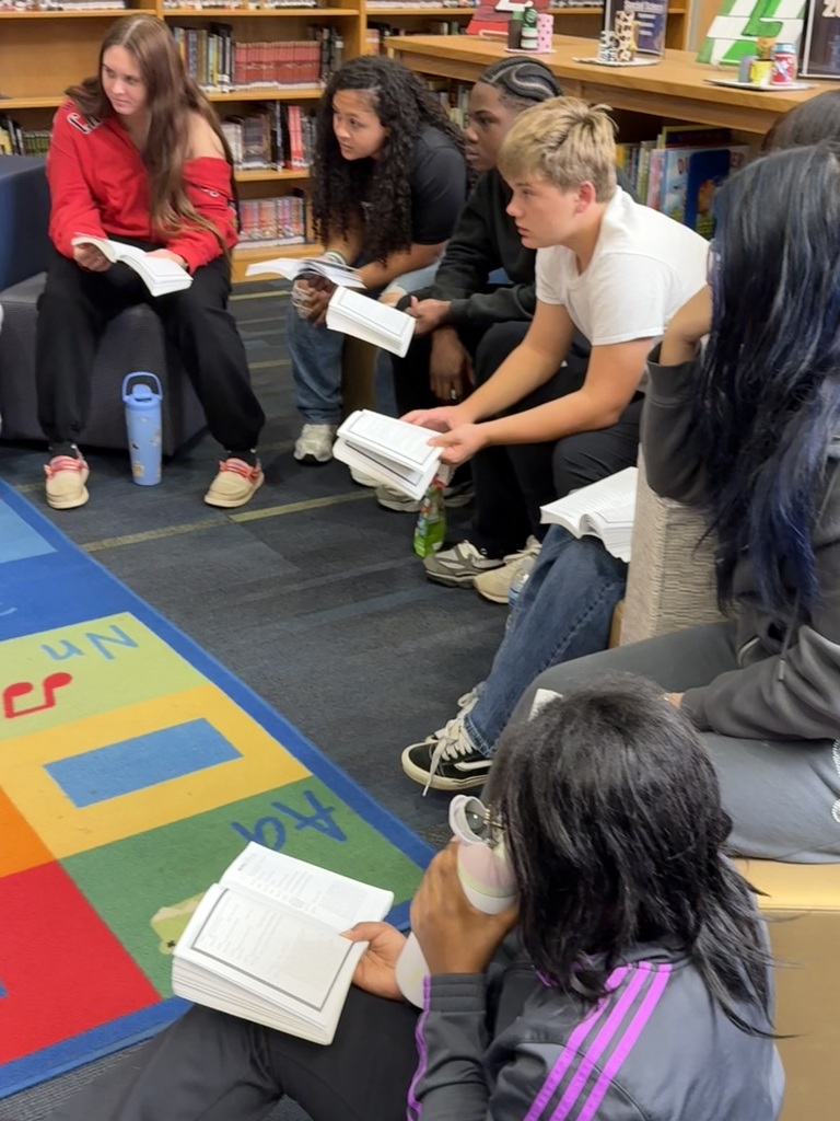 Students sit in a circle on floor cushions, some reading books, with an alphabet rug under them.