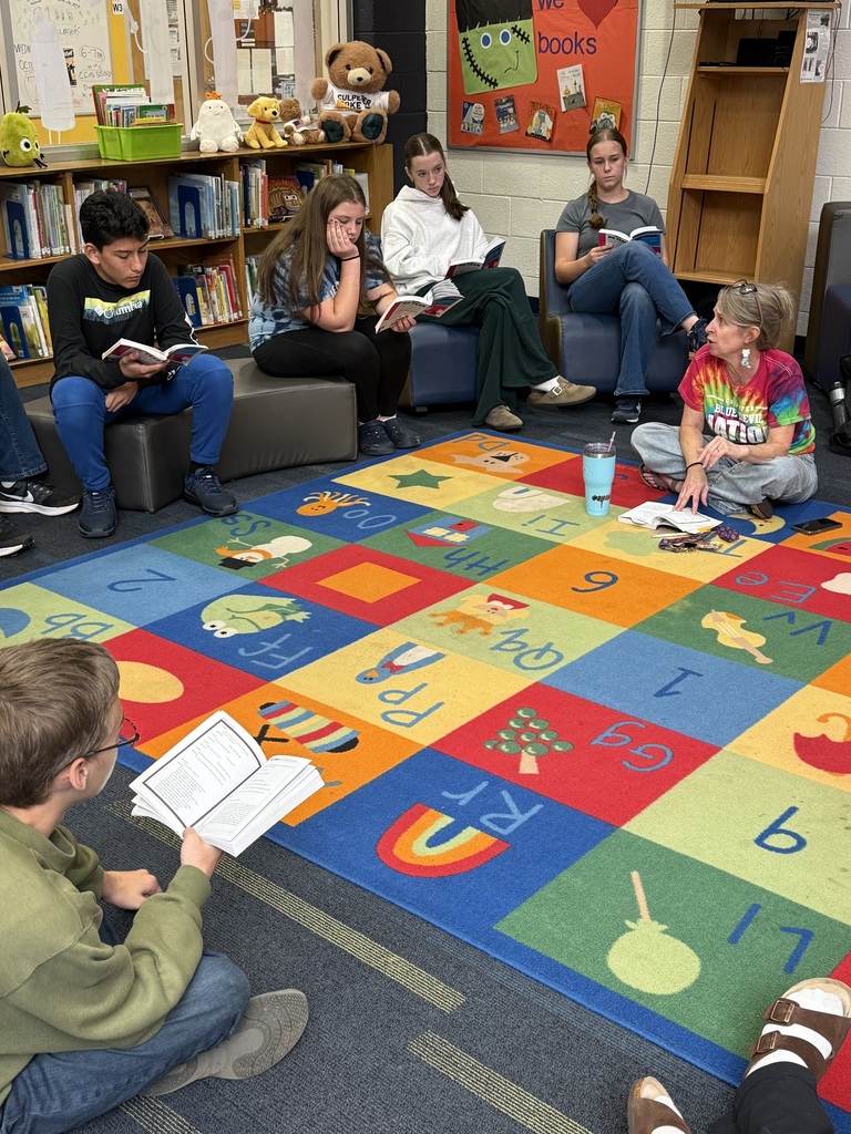 Students sit in a circle on floor cushions, some reading books, with an alphabet rug under them.