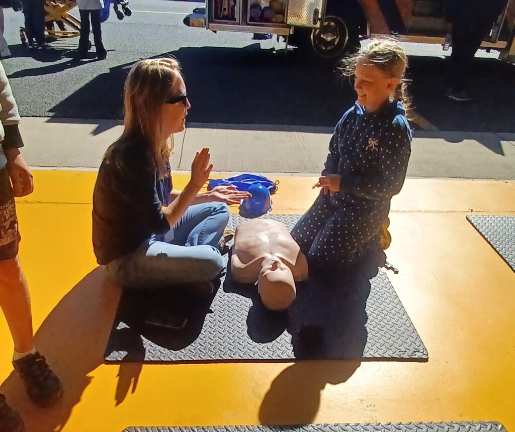 Two women kneeling on mats with a CPR manikin. One woman wearing sunglasses. Bright yellow background.