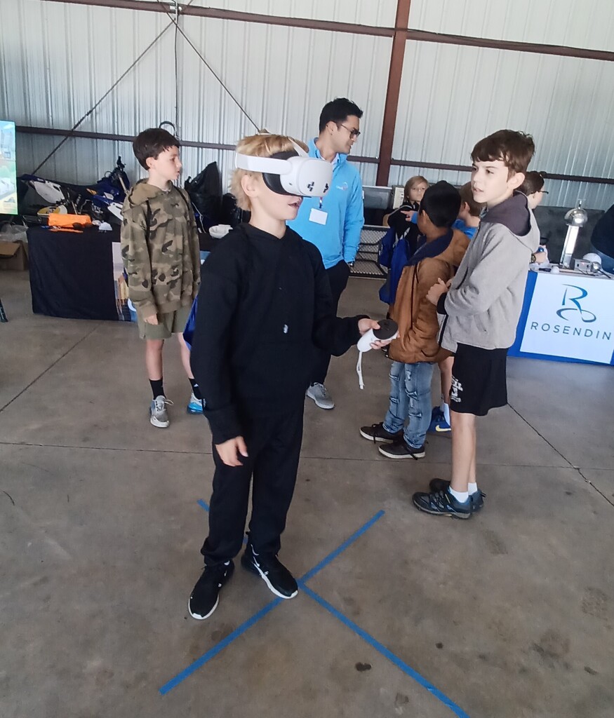 Children wearing VR headsets interact in an indoor area with a table and a sign behind them.
