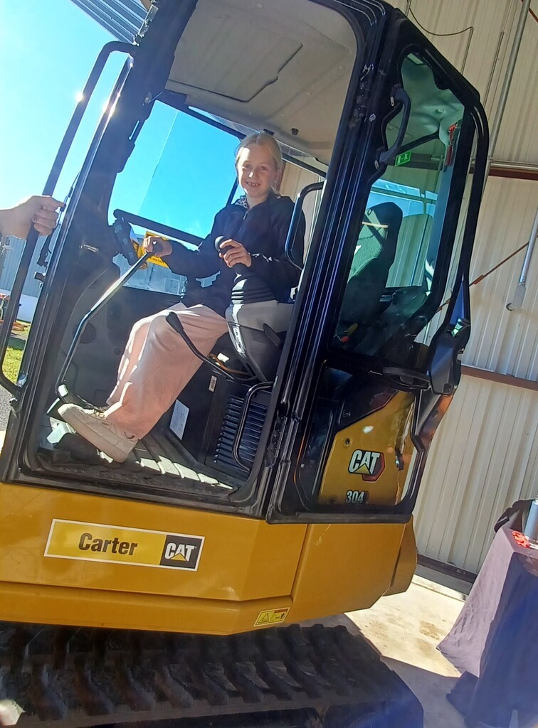 A woman sits in a yellow bulldozer, her legs crossed, wearing a black top and pink pants.