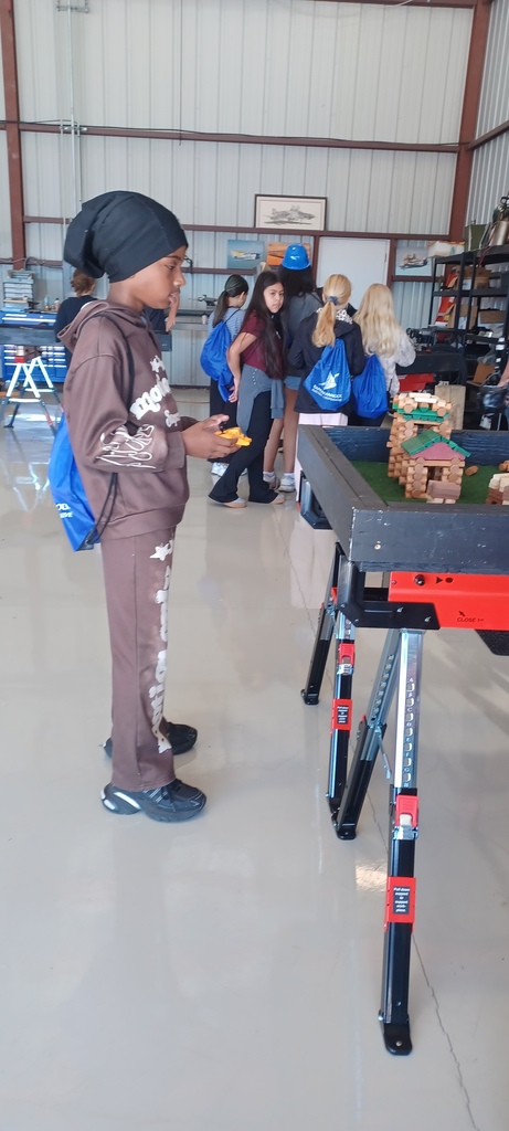 A boy with a blue backpack examines a wooden birdhouse on a table in a workshop.