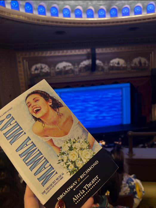 A hand holds a program with a bride image. A large stage is behind with blue lights and a screen.