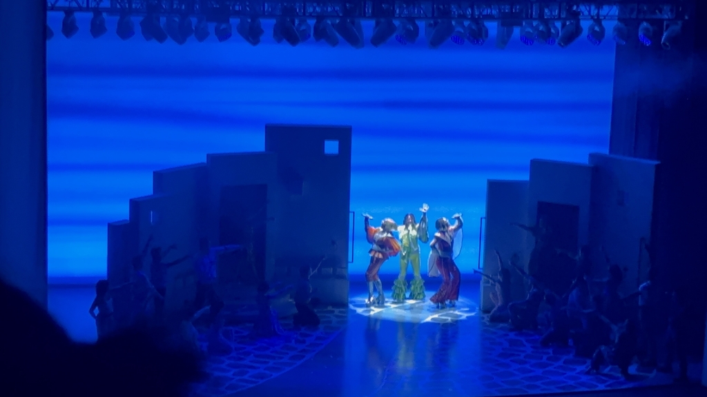 Stage with performers in vibrant costumes under a blue light backdrop, highlighting a central dance.