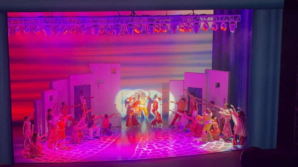 A stage with performers in bright costumes dancing. Purple and pink lights illuminate the scene.