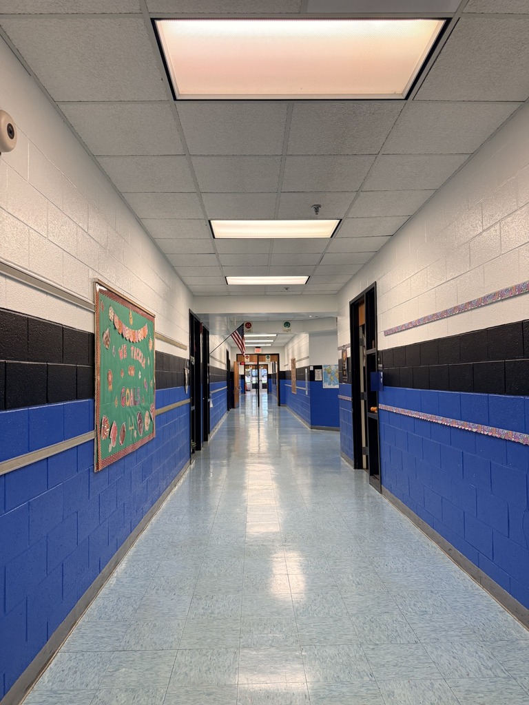 Empty school hallway with blue and white walls, ceiling lights, and a framed poster on the left.