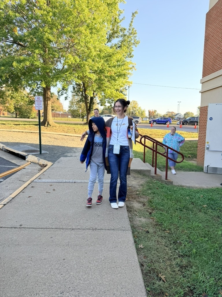 Two individuals walk on a sidewalk, one wearing glasses and a white top, the other in a blue jacket. Trees and a building are in the background.