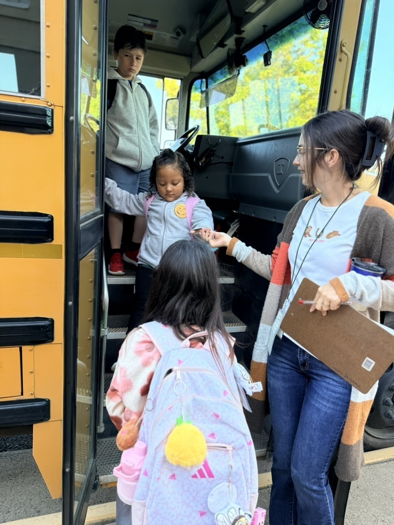 A school bus with three children, one wearing a backpack, and a woman with glasses holding a folder.