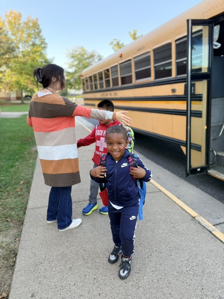 A woman assists two children, one with a backpack, walking toward a school bus on the sidewalk.