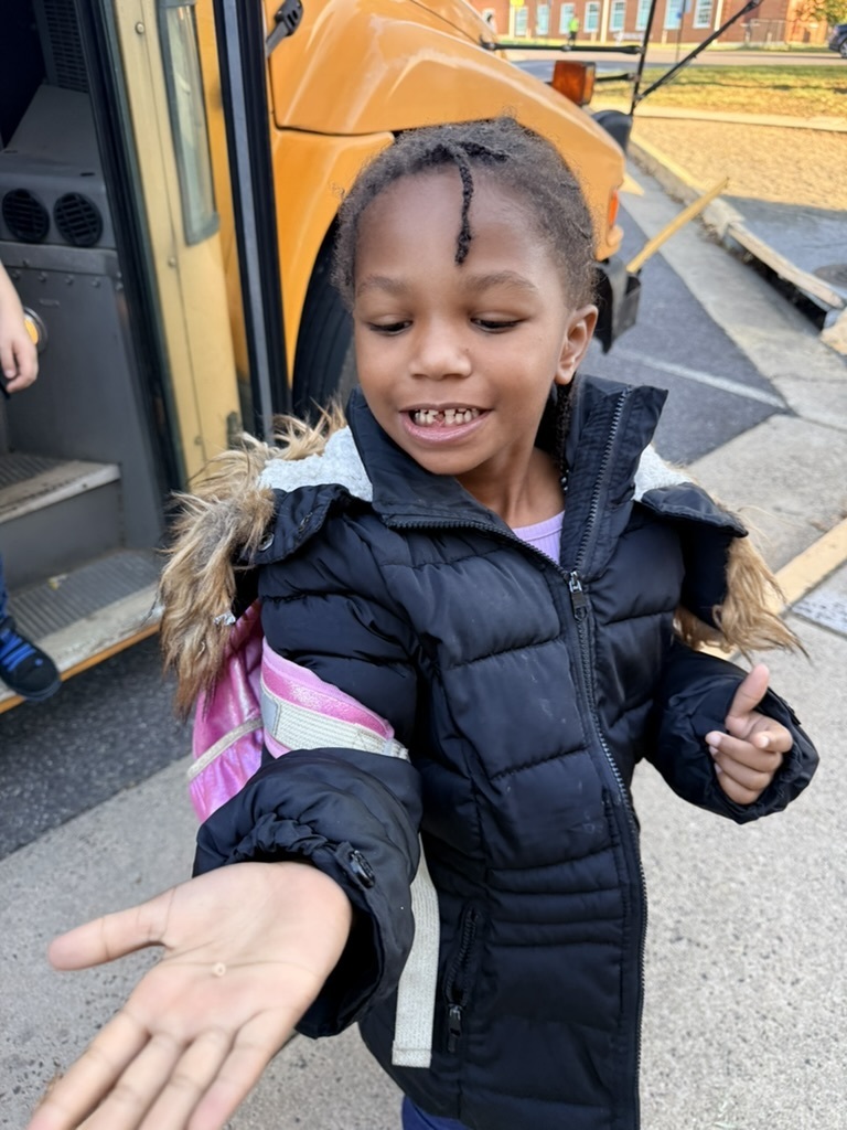 A young girl in a black coat shows a hand with a small object in front of a yellow school bus.