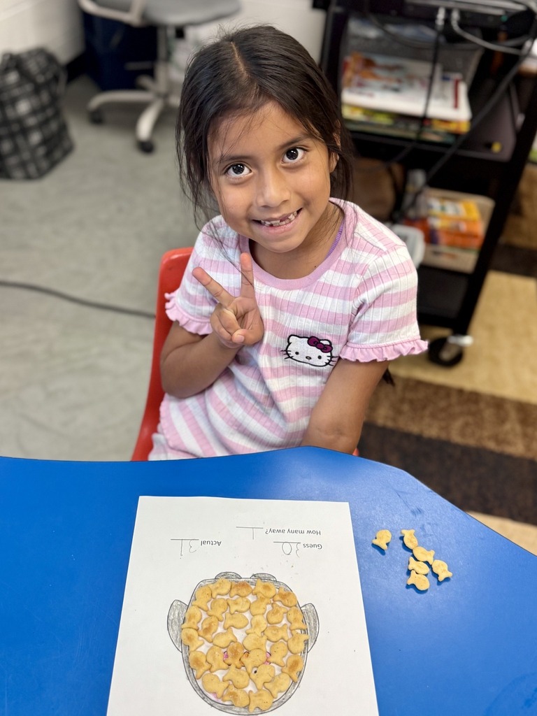 A young girl in a pink shirt sits at a table, smiling and making a peace sign.