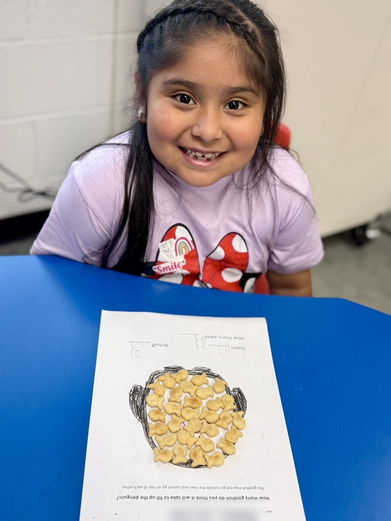 A young girl with braided hair sits at a blue table, smiling, with a paper in front of her. The paper features a drawing of popcorn with accompanying text.