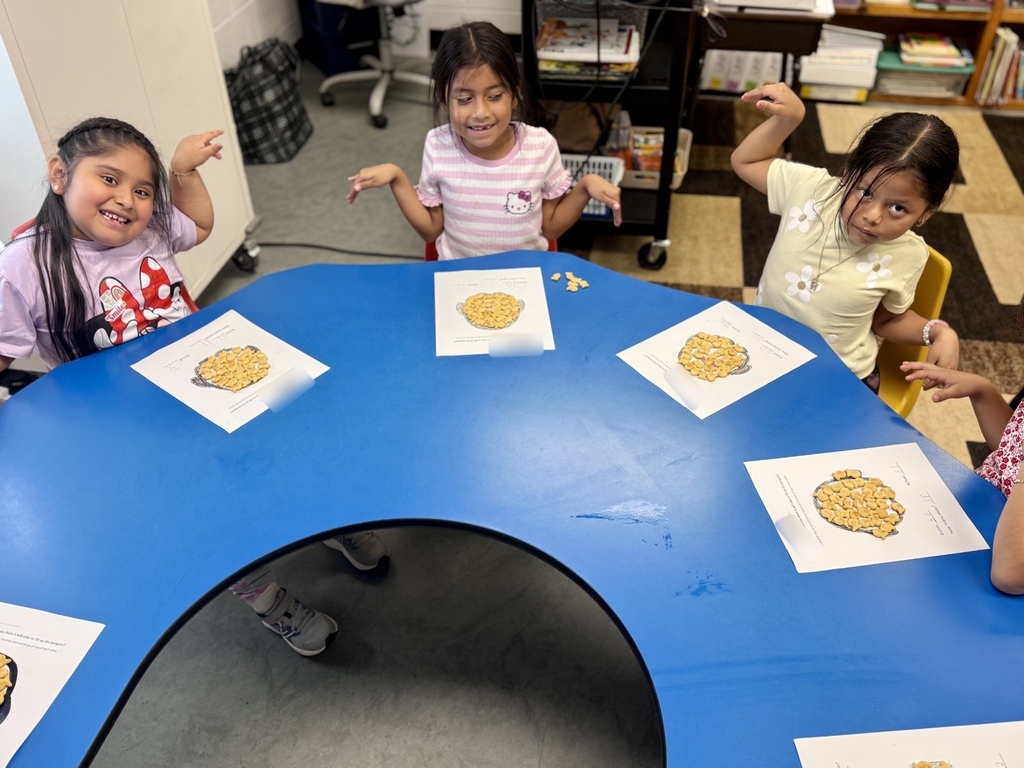 Four children sit at a blue table, looking at pictures of cookies on paper. Shelves with books are in the background.