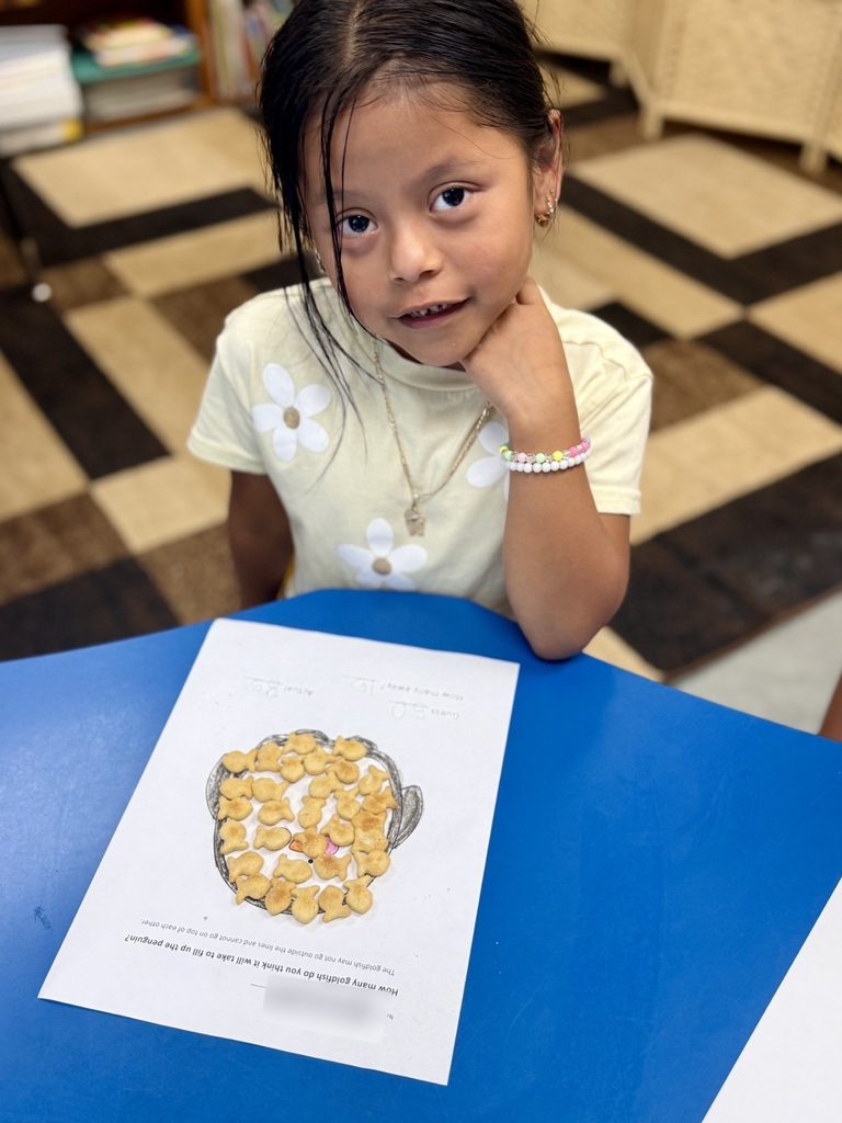 A young girl sits at a blue table, posing with a hand on her chin. A paper with a drawing of popcorn is in front of her.