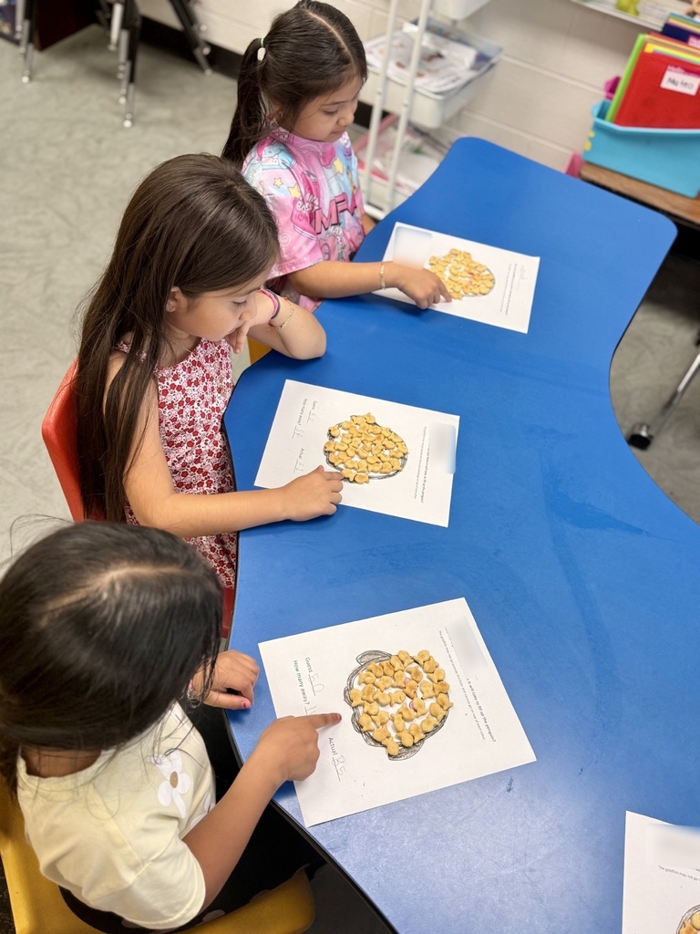 Three young children sit at a blue table, pointing at and interacting with snack foods on white papers.