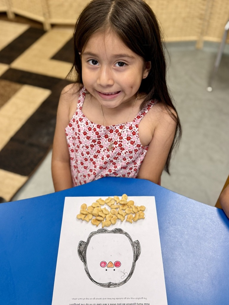 A young girl sits at a blue table, smiling. She has a paper with a drawn face and cereal on it.