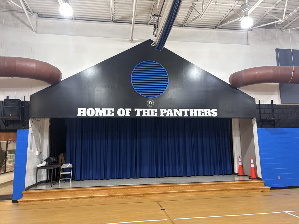 Gymnasium stage with black backdrop. It reads "Home of the Panthers" with blue curtain. Orange cones near stage.