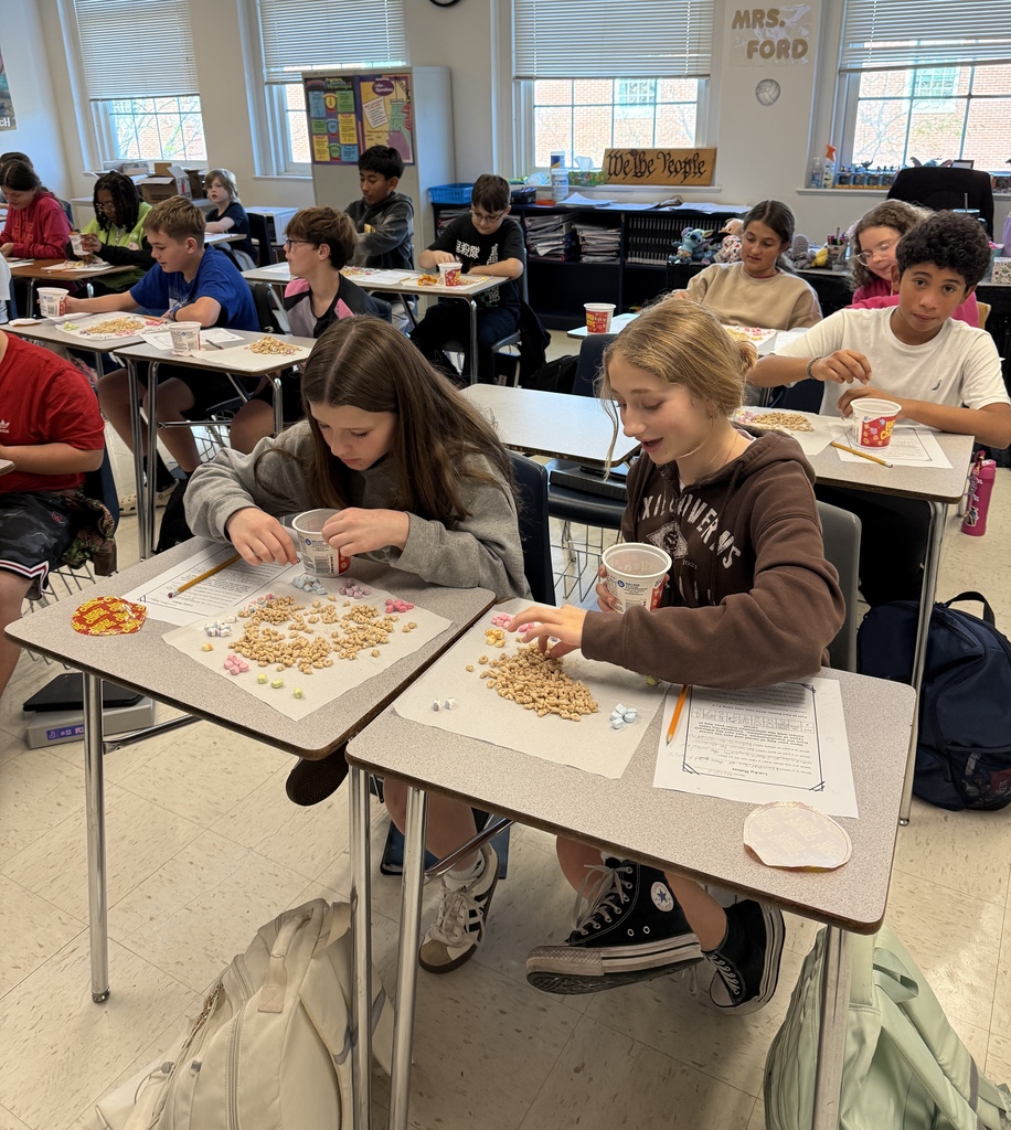 Students in Mrs. Ford’s classroom participate in a hands-on learning activity using colorful cereal and marshmallows. Students sit at their desks sorting, counting, and arranging the cereal and marshmallows on paper towels while following instructions on worksheets. Two students in the foreground carefully organize their materials, while classmates throughout the room work on the same activity.