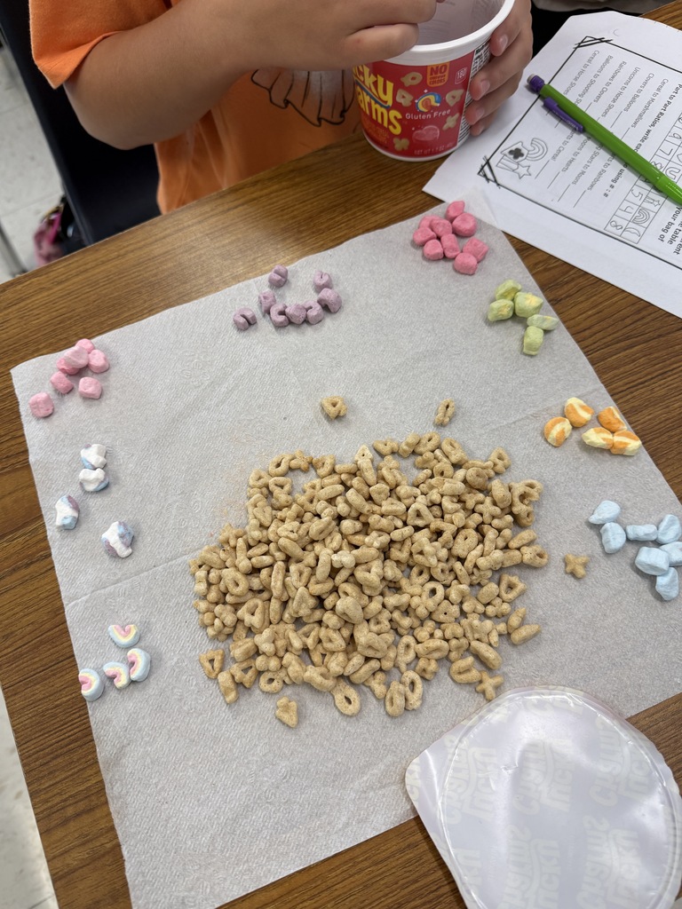 Close-up of a student's Lucky Charms math activity showing a pile of cereal surrounded by colorful marshmallows sorted into groups by shape and color. A worksheet about ratios and a cup of cereal are visible on the desk.