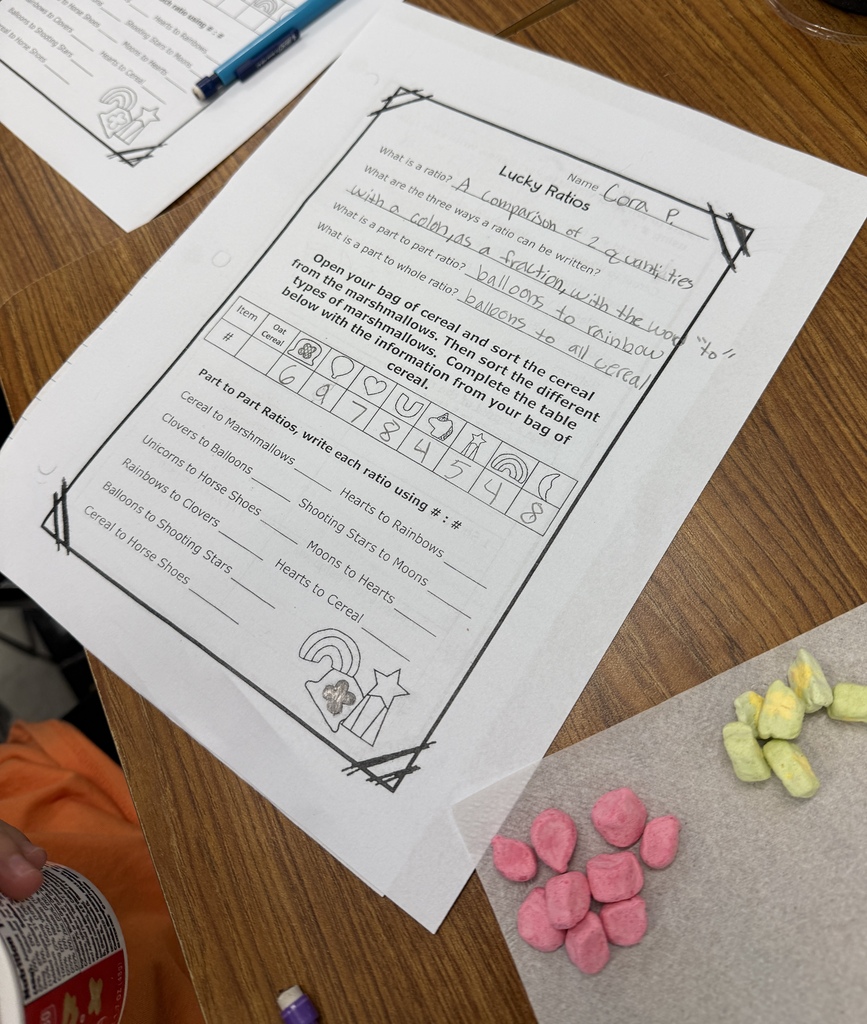 Close-up of a math worksheet titled “Lucky Ratios” showing instructions for sorting Lucky Charms cereal and calculating part-to-part ratios. The worksheet includes a table for recording different marshmallow shapes and their quantities, with colorful marshmallows visible beside it.