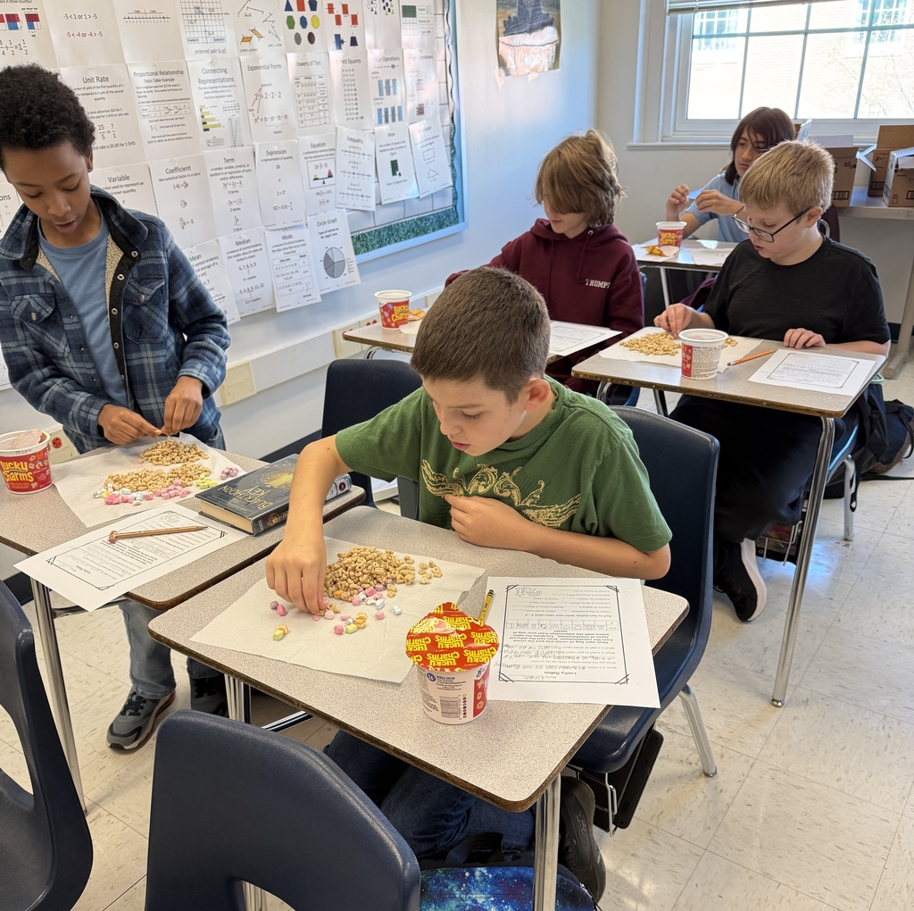 Students in Mrs. Ford’s math classroom participate in a hands-on lesson about ratios using Lucky Charms cereal. Students sort marshmallows into groups, count cereal pieces, and record their findings on worksheets. Math vocabulary posters are displayed on the classroom wall.