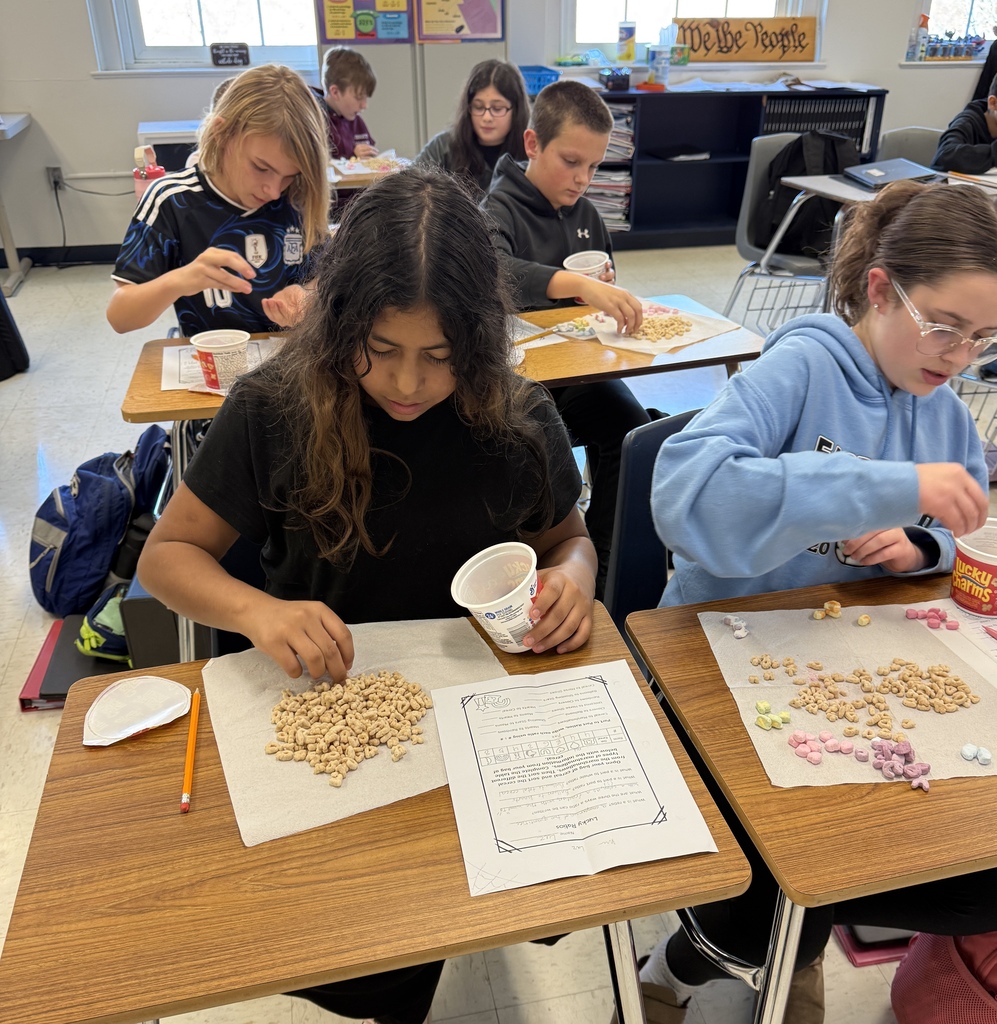 Students in Mrs. Ford’s classroom sort Lucky Charms cereal and colorful marshmallows into groups while completing math worksheets. Two students in the foreground carefully separate cereal pieces by shape and color as classmates work at their desks behind them.