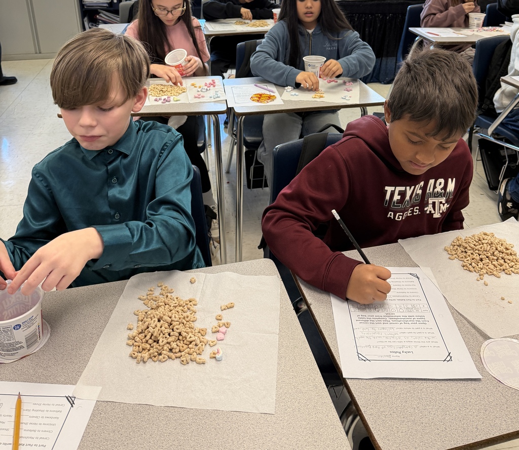 Two students in Mrs. Ford’s classroom work on a hands-on math activity using Lucky Charms cereal. One student sorts cereal and colorful marshmallows while another records answers on a worksheet about ratios. Other students work on the same activity in the background.