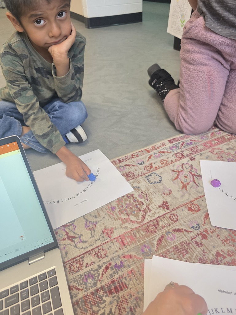 Student sits cross-legged on the classroom floor, holding a blue marker on an alphabet arc worksheet beside a laptop.