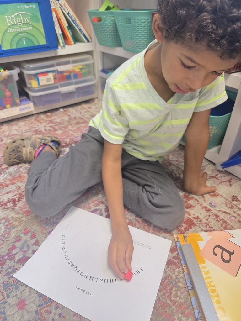 Student in a striped shirt kneels on a classroom rug, placing a red marker on an alphabet arc worksheet.