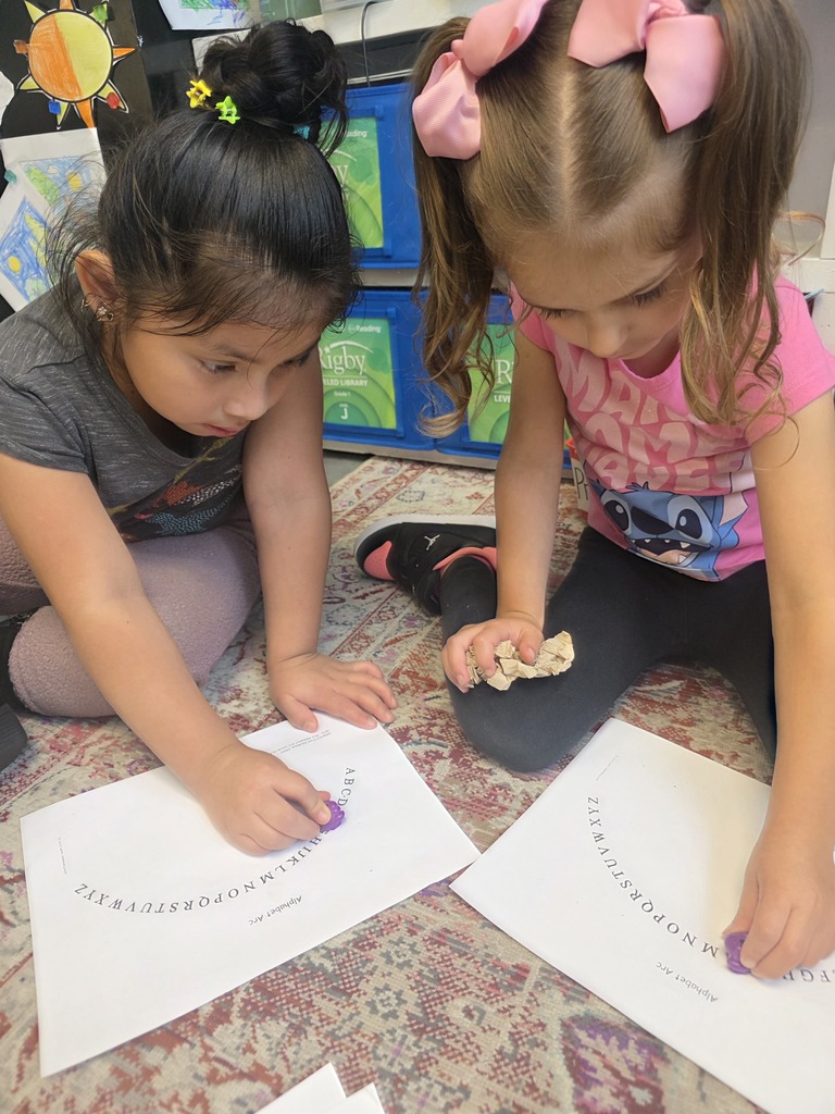 Two students sit together on a classroom rug, placing purple markers along letters on their alphabet arc worksheets.