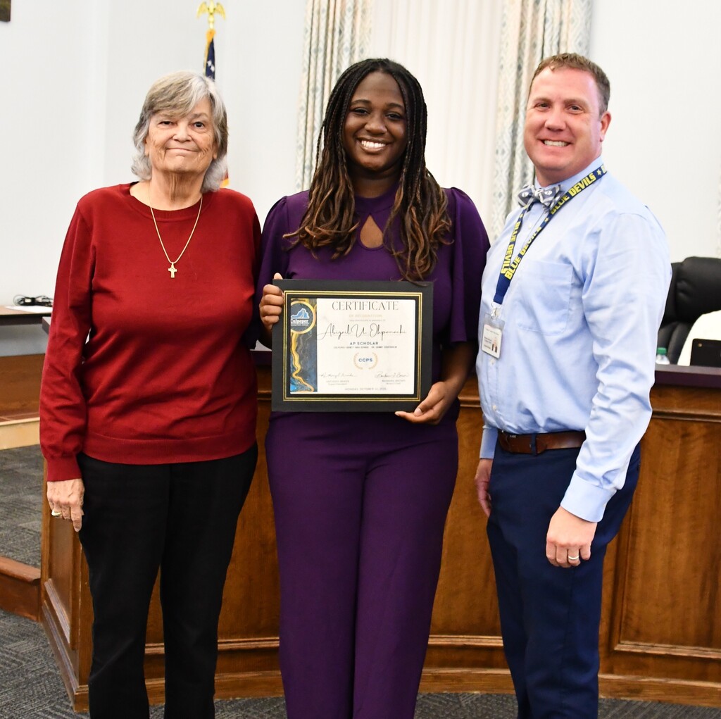 Three people stand in front of a wooden desk. The woman in the middle holds a framed certificate.