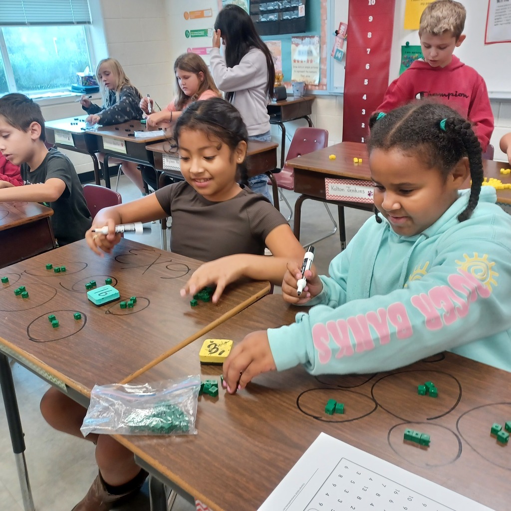 Two girls sit at a table with green blocks and a worksheet, focused on an activity.