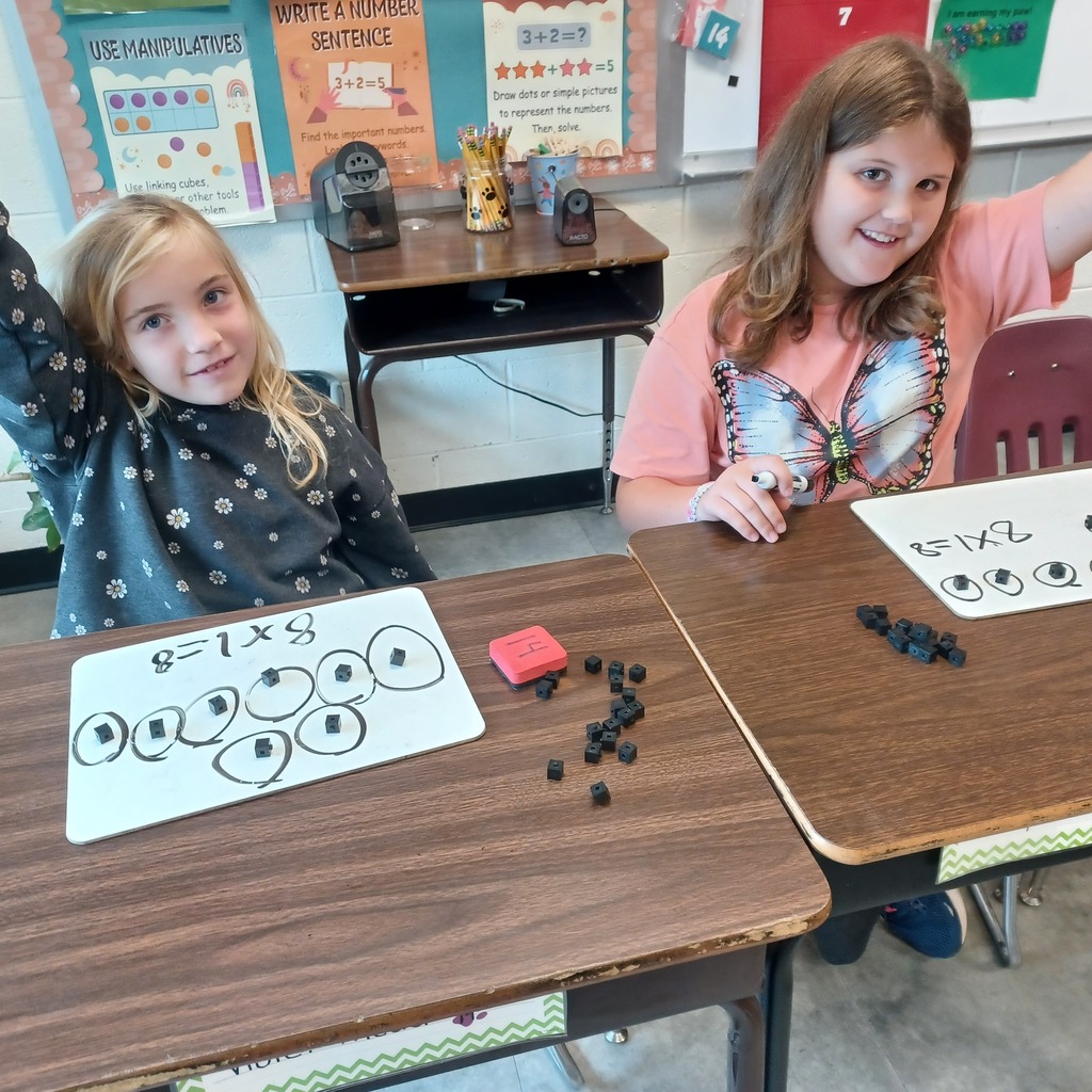Two young girls seated at desks in a classroom. One girl raises her hand; the other holds a paper with a multiplication problem.