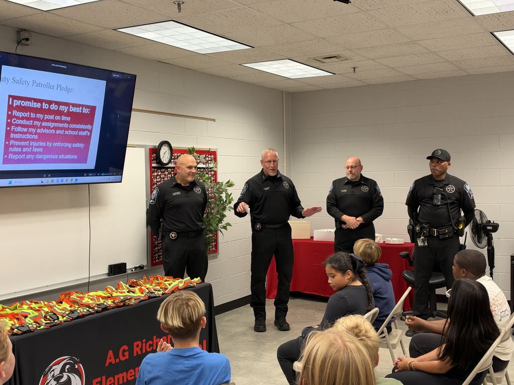 Four officers stand before a crowd, one holding a plant. Monitor on wall displays text. Audience seated in chairs.