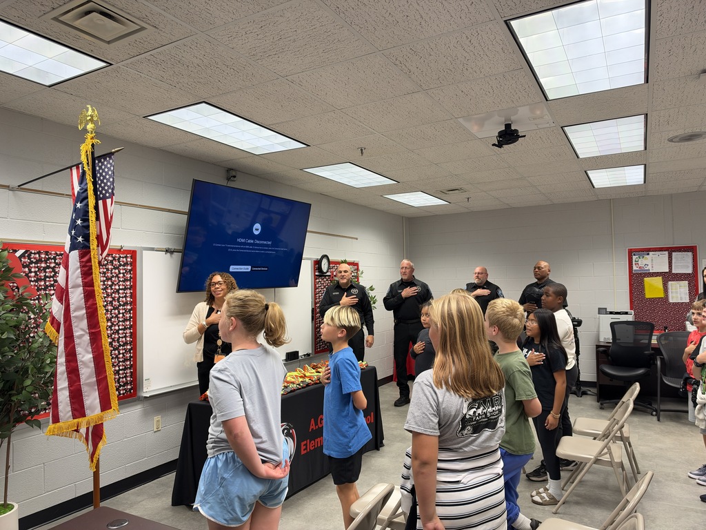 A classroom setting with children standing in a circle, one woman speaking, a screen on the wall, and an American flag on a pole.