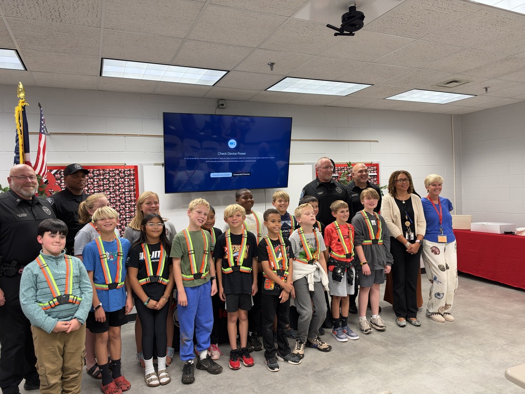 A group of children in safety vests stands in a room with a large screen. An adult stands behind them.