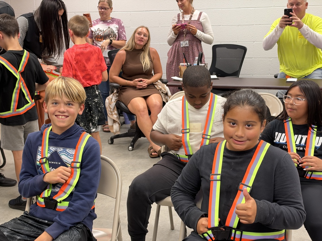 Group of kids and adults wearing safety vests sit in chairs in a room. One woman takes a photo.