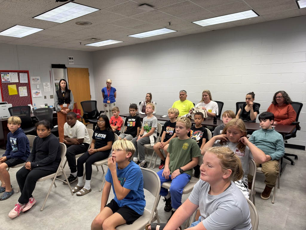 In a room with white walls and ceiling lights, children and adults sit in chairs, listening to a person standing.