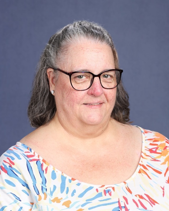 An adult woman with gray hair and glasses smiles against a blue background. She wears a colorful blouse.