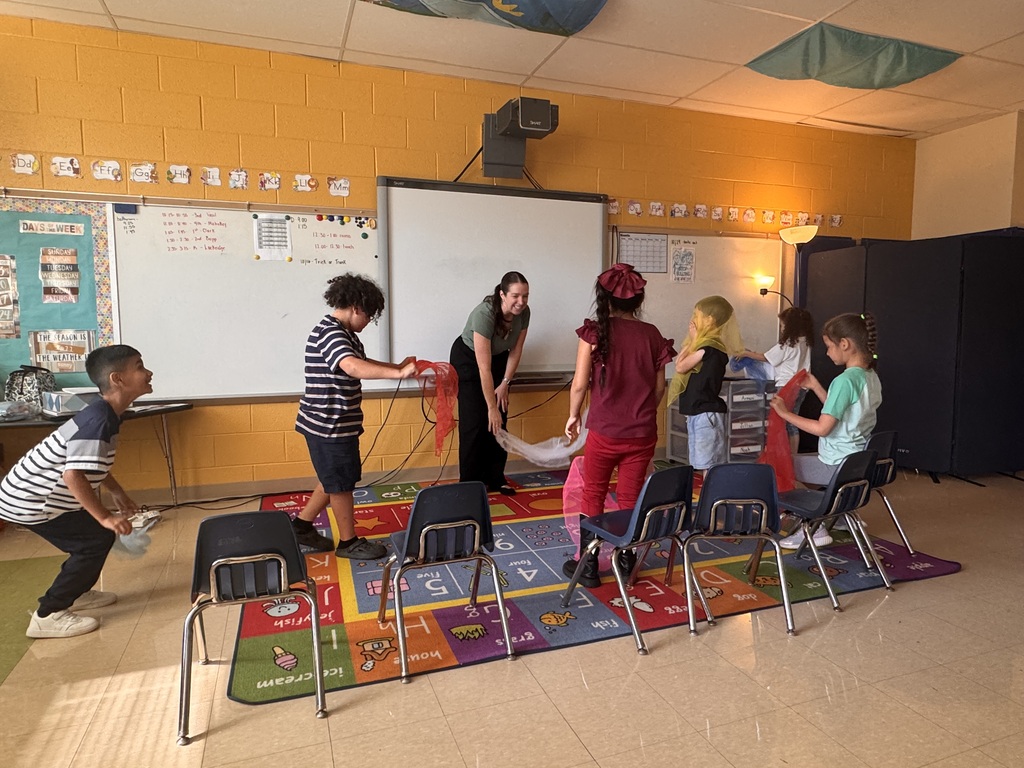 Children and two adults in a classroom. They stand around a rug with letters, numbers, and pictures.