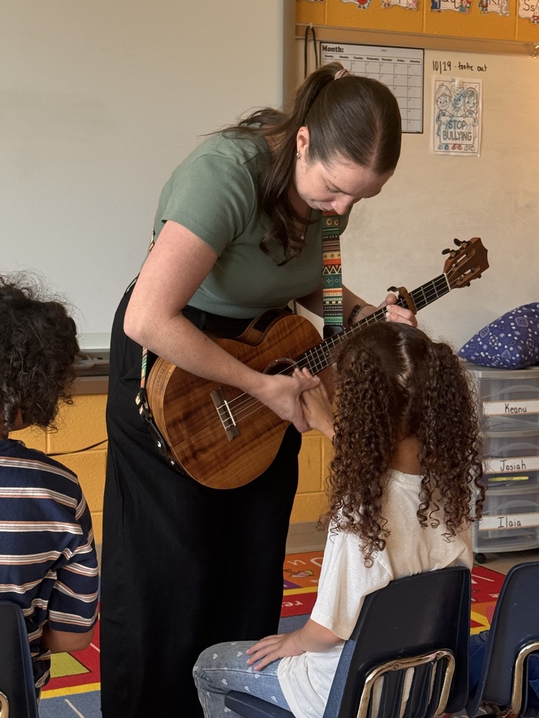 An adult stands and plays a ukulele while a child sits in a chair. Two other children sit behind.