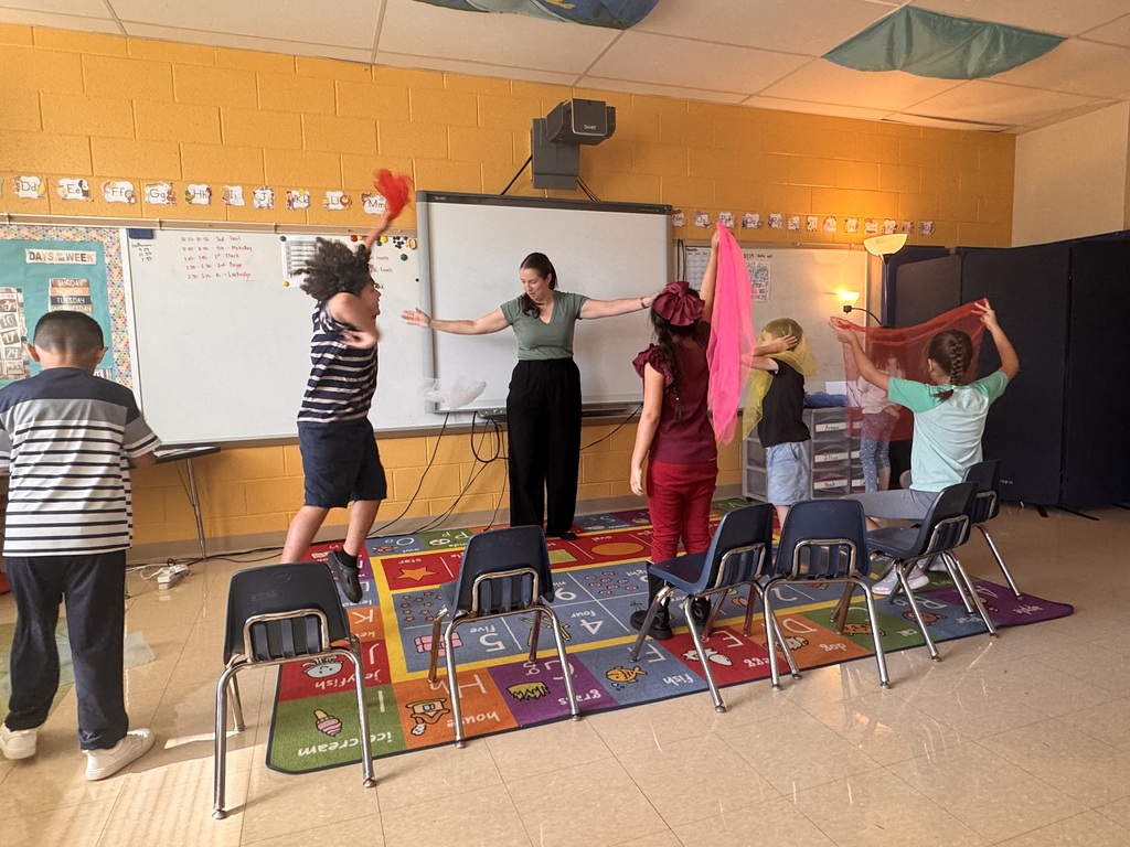 Group of children and adults in a classroom. Some stand with scarves while others sit. A whiteboard is on the wall.