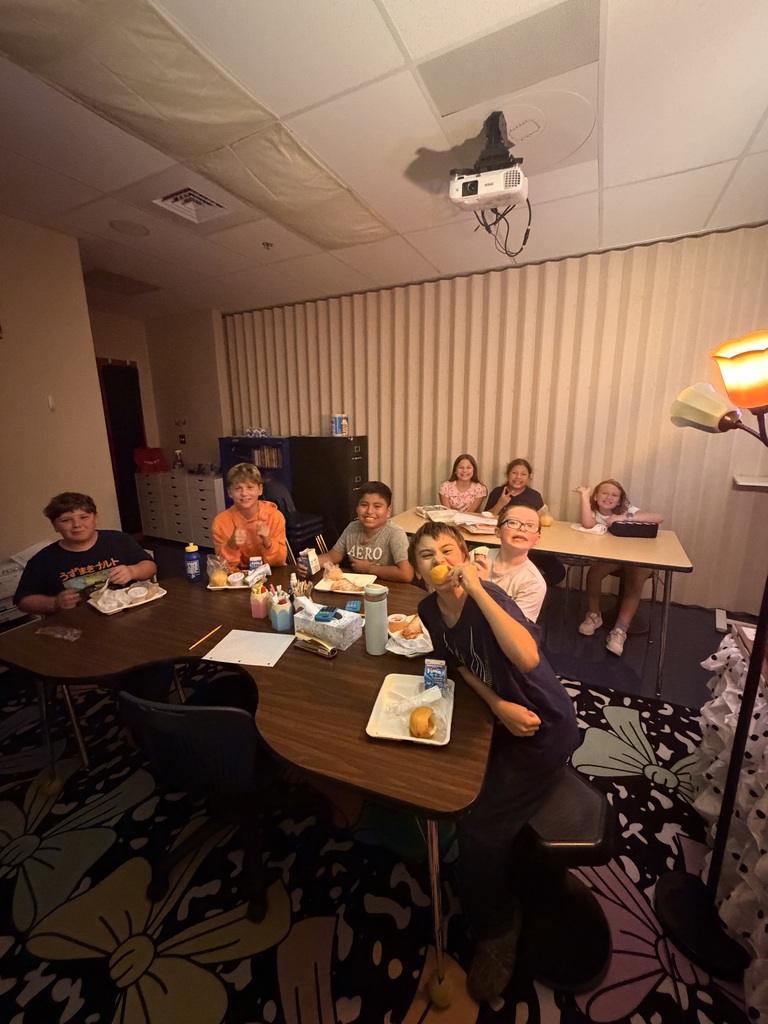 Several children sit around a table in a room. Some eat with their hands while others hold utensils.